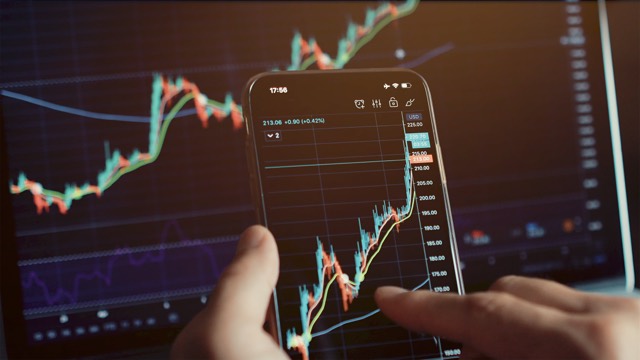 Checking a rising chart on a phone in front of trading screens